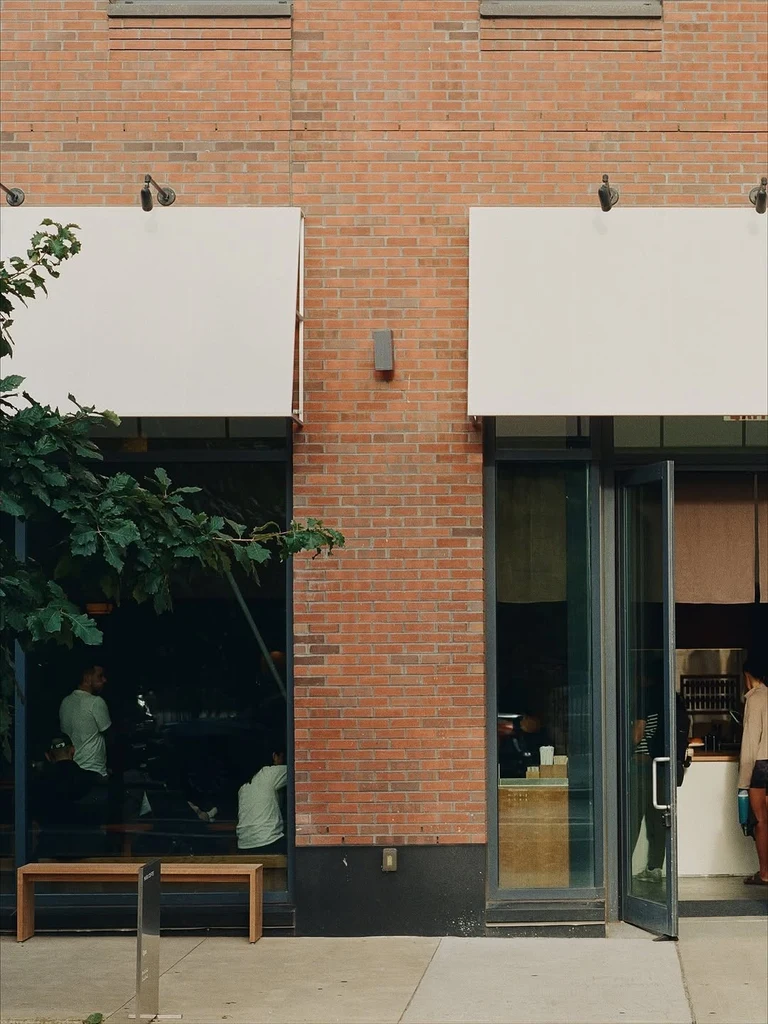 Brick cafe front with two plain cream canvas awnings over its windows and open glass door