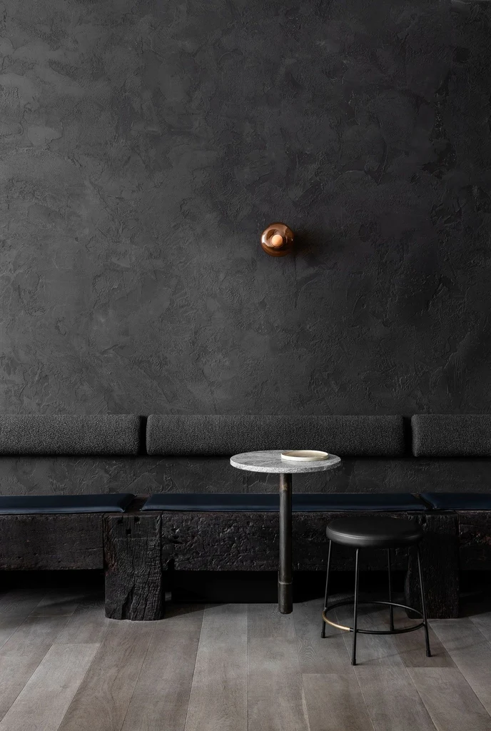 Charcoal plaster restaurant wall with a copper wall light, a banquette, a stool and a stone table