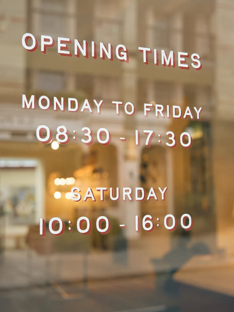 Stejná výloha s hodinami změněnými na 08:30 - 17:30 ve všední dny a 10:00 - 16:00 v sobotu stejným bílým písmem s červeným stínem; nadpisy i interiér za sklem zůstaly beze změny.