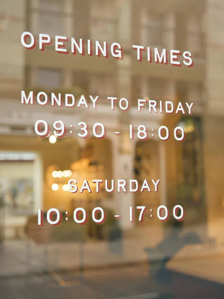 Fotka výlohy obchodu s bílým vinylovým nápisem OPENING TIMES, MONDAY TO FRIDAY 09:30 - 18:00 a SATURDAY 10:00 - 17:00 s červeným posunutým stínem na pozadí jemně rozostřeného interiéru.