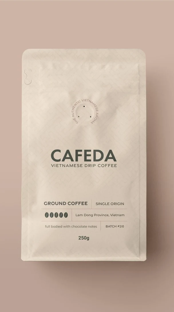 くすんだピンクの背景に置かれた、CAFEDA のベトナム式ドリップコーヒーのクリーム色のスタンドパウチ。ラベルには「ground coffee」「single origin from Lam Dong Province」「full bodied with chocolate notes」「batch #210」「250g」と書かれている。