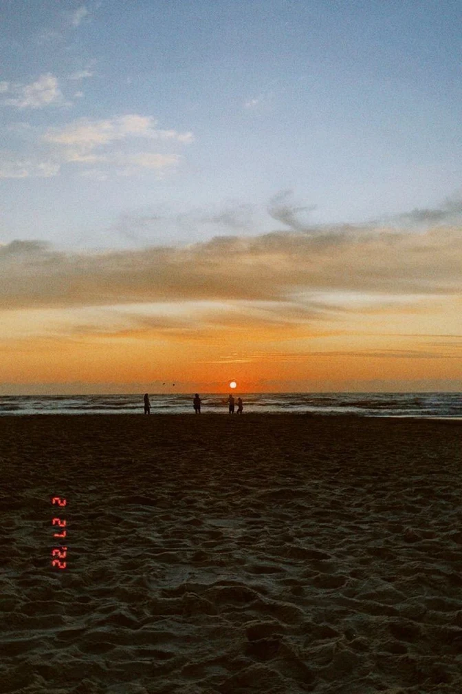 Film photo of a beach at sunset with small silhouettes at the waterline and an orange camera date stamp reading 2 27 ’22 printed sideways over the sand near the lower left edge.