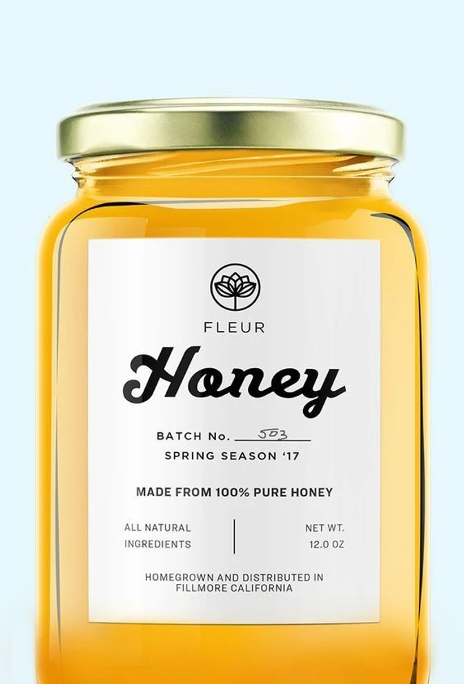 装有蜂蜜的玻璃罐，贴着白色 FLEUR 标签，上面用黑色手写体写着 Honey，还有手写批号 503、“spring season ’17”、“made from 100% pure honey”、“all natural ingredients”、“net wt. 12.0 oz”以及“homegrown and distributed in Fillmore California”